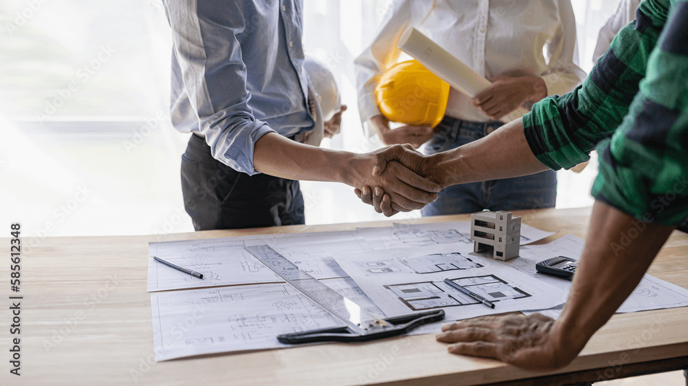 Three men stand over a table covered in diagrams. Two of the men are shaking hands across the table.