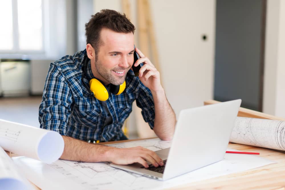 A man sits on his cellphone in front of a laptop. He has noise cancellling headphones around his neck while he works.