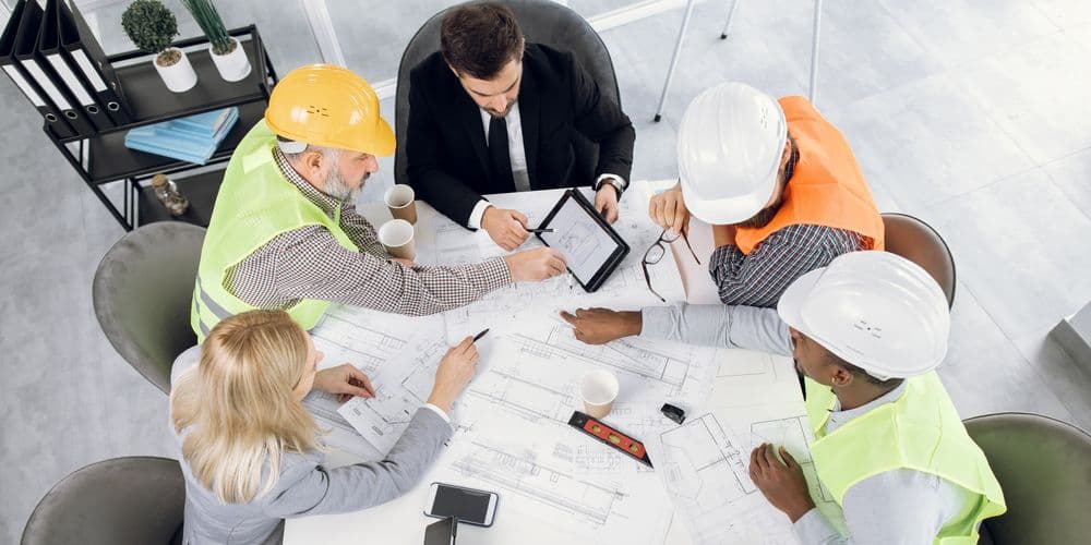 One woman and four men sit around a table looking at a tablet. There is various document scattered on the table. Three men are wearing hard hats and hi-vis vests.