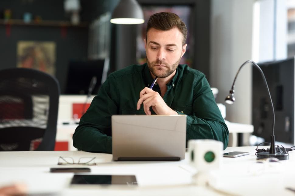 A man sits looking at his laptop. He holds a pen in his hand and looks to be in thought.