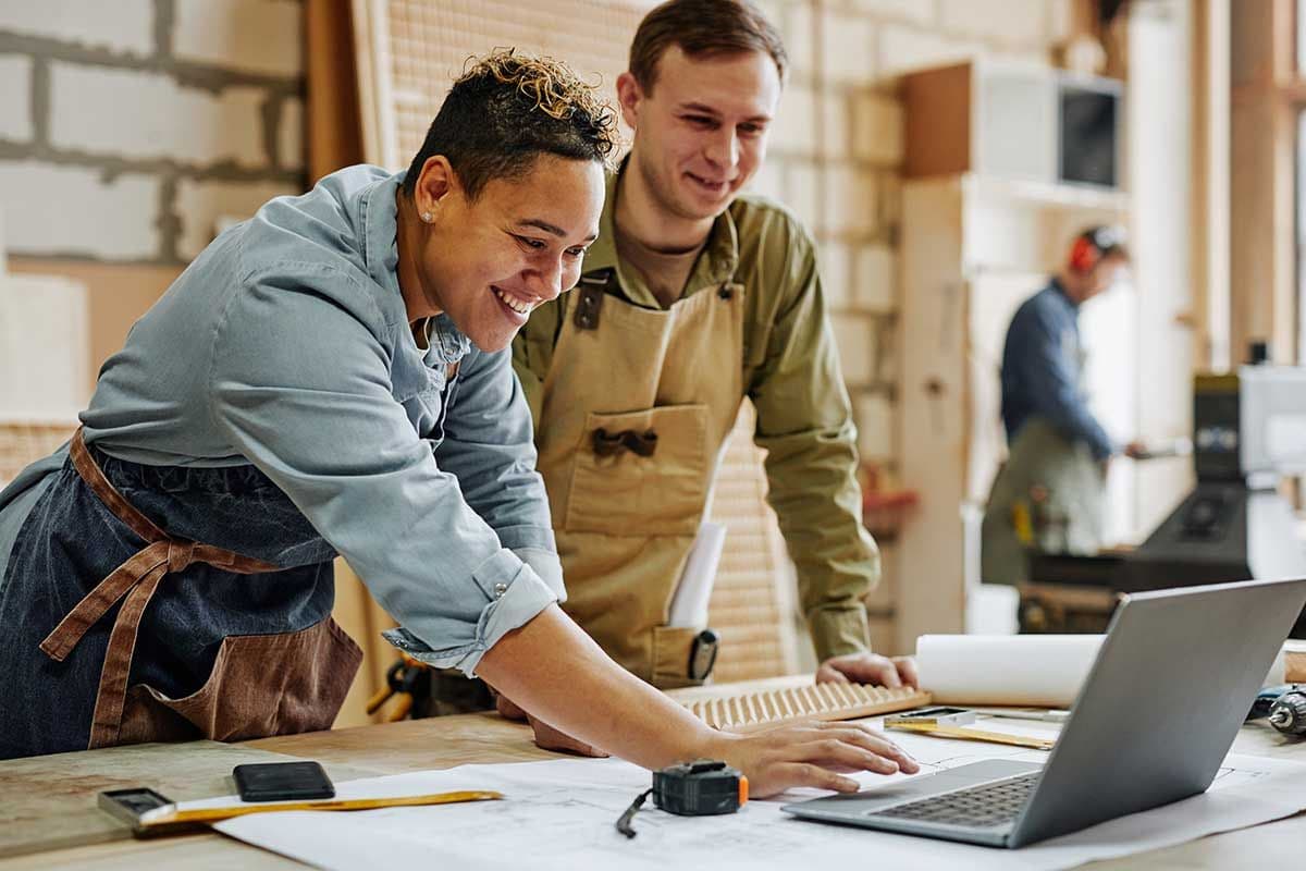 Two people stand in front of a laptop. They both wear aprons like they are working with tools. The person on the left is using the trackpad of the laptop while the other person smiles and watches.
